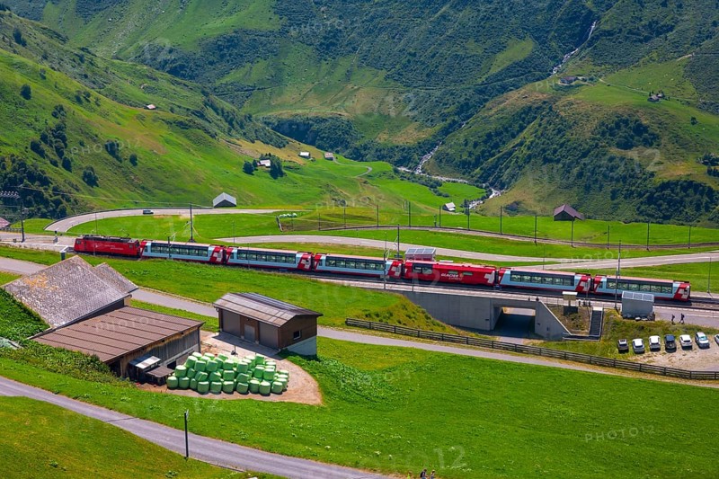 Beautiful View over Mountain Valley with Mountain Road and Railroad Station with Glacier Express Train in a Sunny Summer Day in the Swiss Alps in Andermatt, Uri, Switzerland