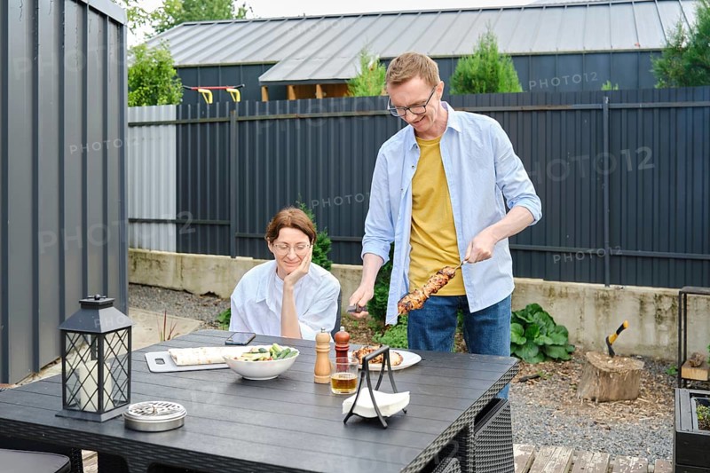 Middle-aged couple enjoy a barbecue meal outdoors in a backyard. Man is serving skewers on a grill, while his wife watches happily. The setting is sunny, with plants and outdoor furniture around