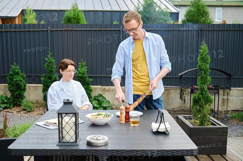 A man is serving grilled meat to a woman seated at a dining table in a beautiful backyard. The garden is well-maintained, and a fresh salad is present. It is a warm, sunny day