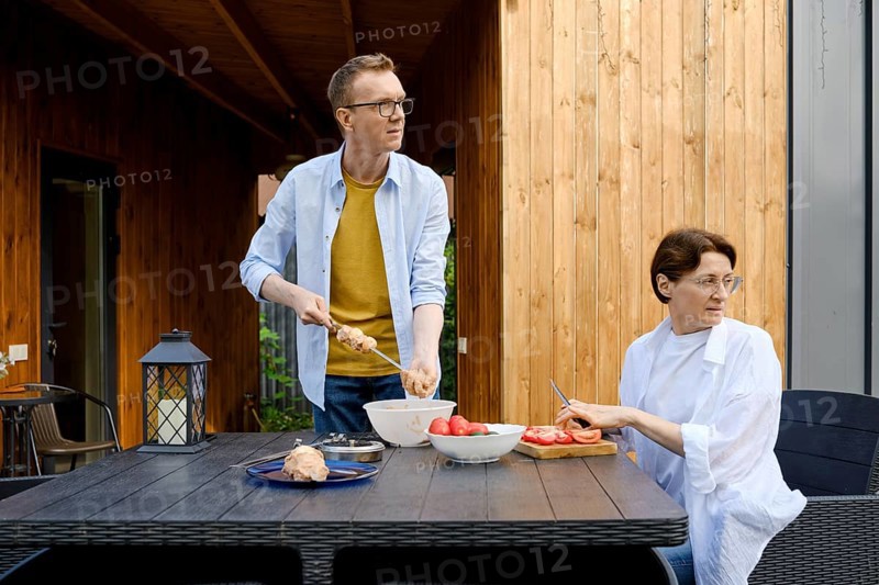 During a sunny day a couple is engaged in preparing a meal outdoors. One person is skewering food while the other slices tomatoes, both are distracted from their work and look away