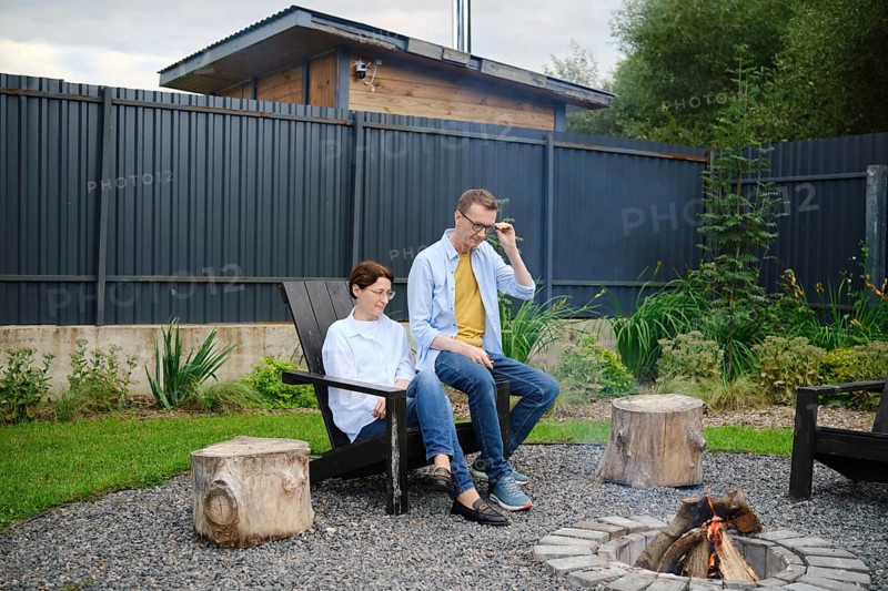 Two people enjoy a tranquil moment by the fire pit in a lush backyard. They share stories and meditatively watching the fire