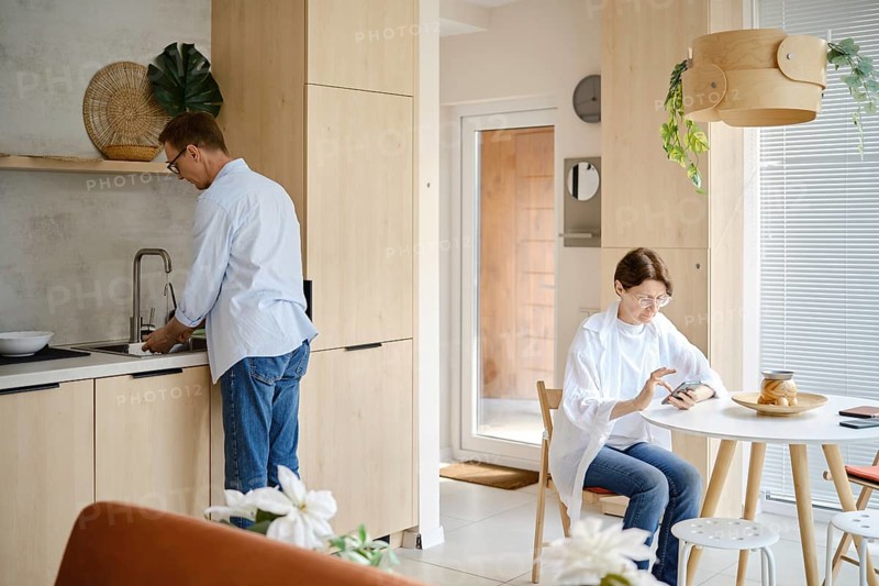 In a contemporary home, a man washes vegetables at the sink while a woman sits at a table, engaged with her phone. Bright, natural light fills the space, highlighting the modern design and warm ambiance