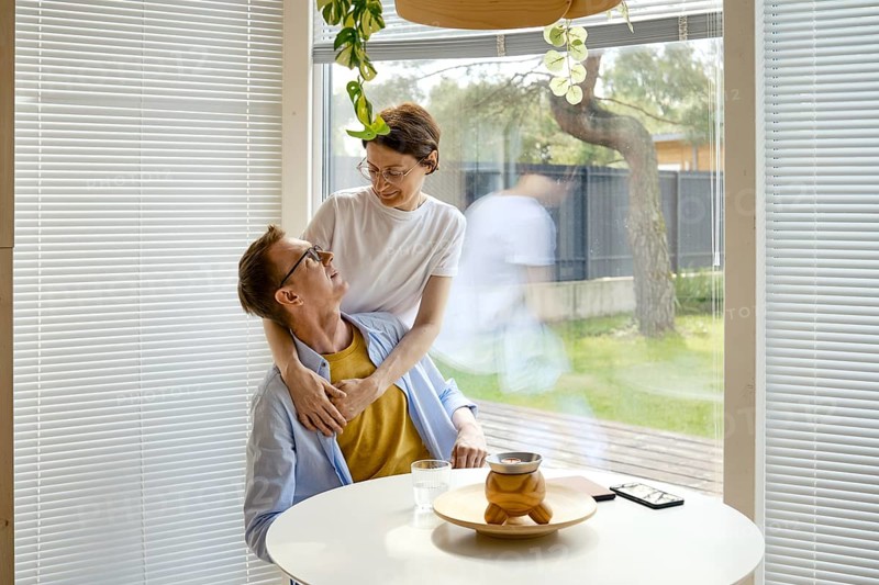 A couple shares a joyful interaction at a modern kitchen table. The woman embraces the man from behind while they engage in conversation, surrounded by natural light and green plants