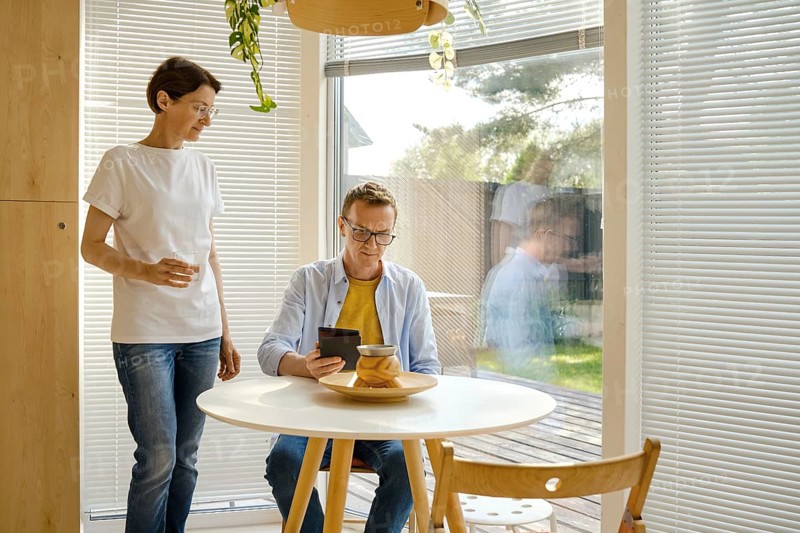 Two people relax in a modern indoor space filled with natural light. One person stands nearby holding a beverage, while the other sits at a table, focused on an e-book