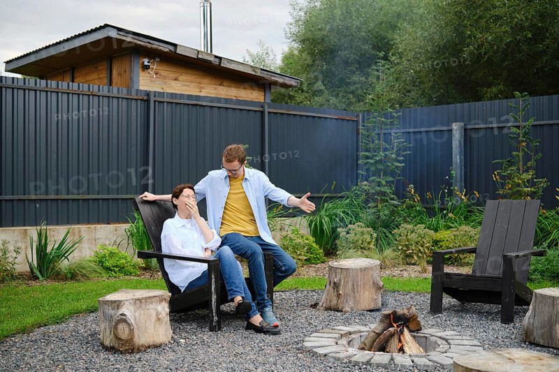 A couple spends a relaxed evening outdoors near a fire pit in their backyard. They share playful banter, surrounded by lush greenery and wooden seating, creating a warm and inviting atmosphere