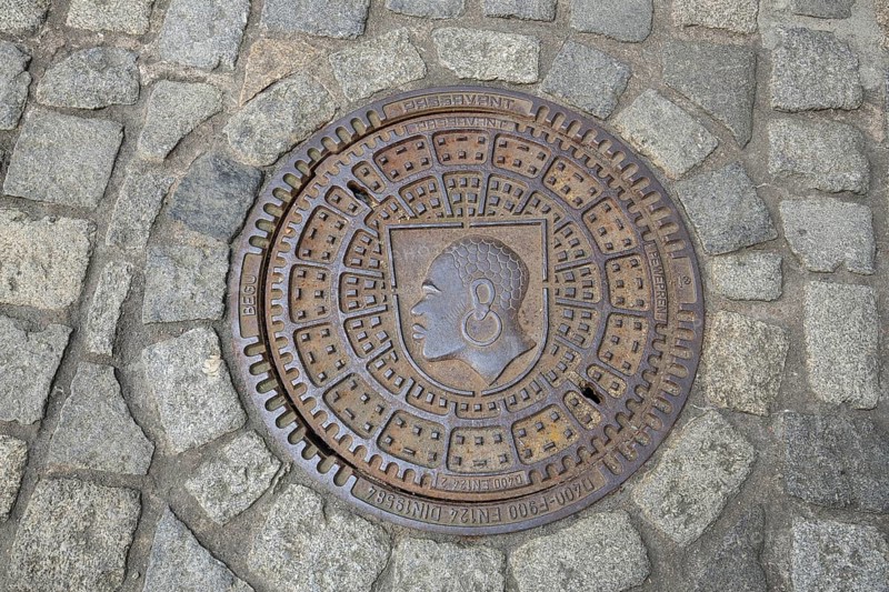 Round manhole cover with city coat of arms, inscription, cobblestones, floor, head, African, earring, Coburg, Upper Franconia, Franconia, Bavaria, Germany