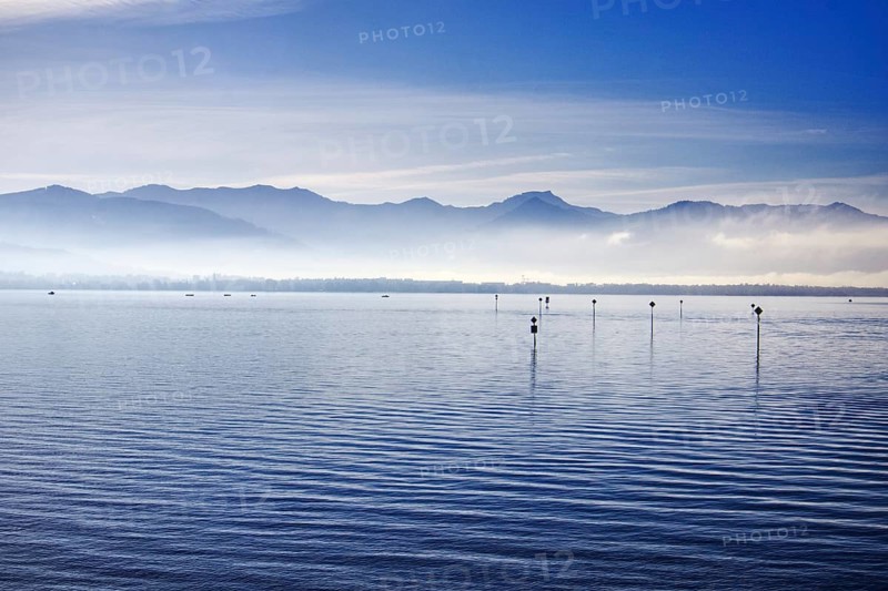 A calm Lake Constance with mountains and fog in the morning light under a blue sky, Lindau