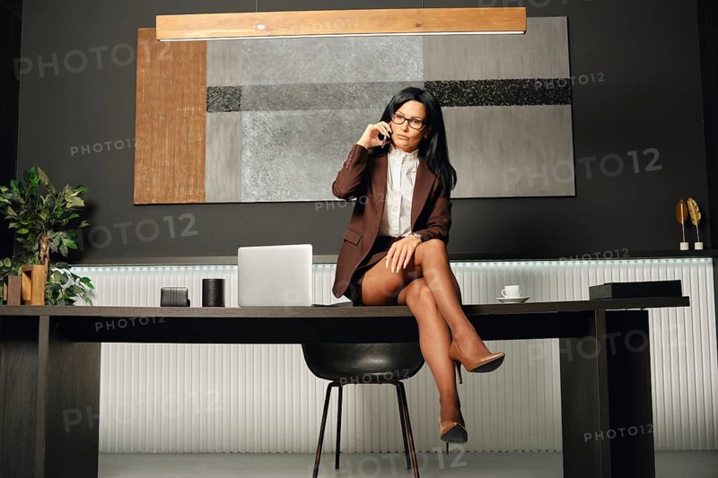 A professional woman in a brown suit sits on a desk while talking on her phone in a sleek, modern office. The dark walls are adorned with abstract art, creating a sophisticated atmosphere