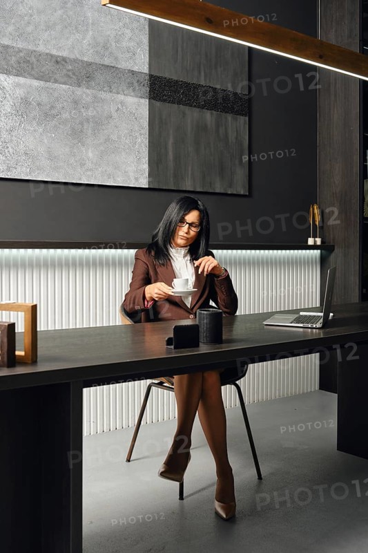 A woman in business attire sits at a sleek desk in a modern office, taking a cup of coffee. The setting features elegant decor and a laptop, creating an atmosphere of productivity and style