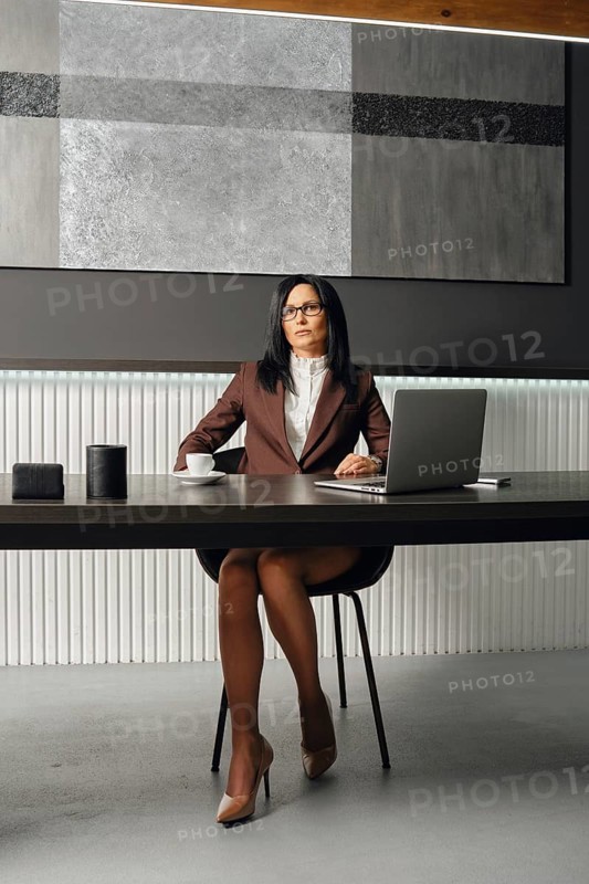 A slender woman with glasses and a stylish suit sits at an office desk with opened laptop. She stares sternly at the camera