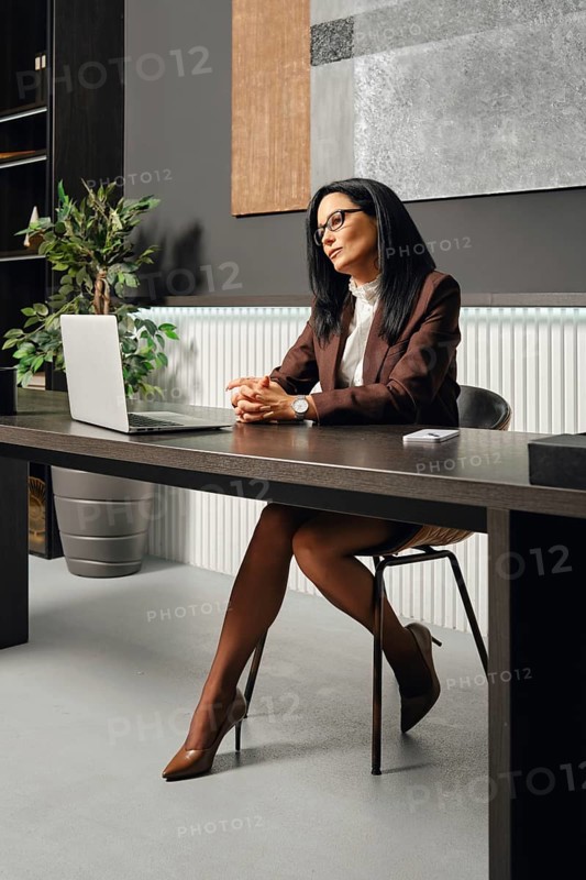 A woman dressed in business attire sits at an office desk in office and looking at the screen of laptop during a video call