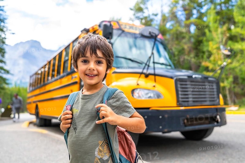 Happy schoolboy carrying backpack is waiting for the school bus in front of blurred mountain landscape, ready to start a new educational year