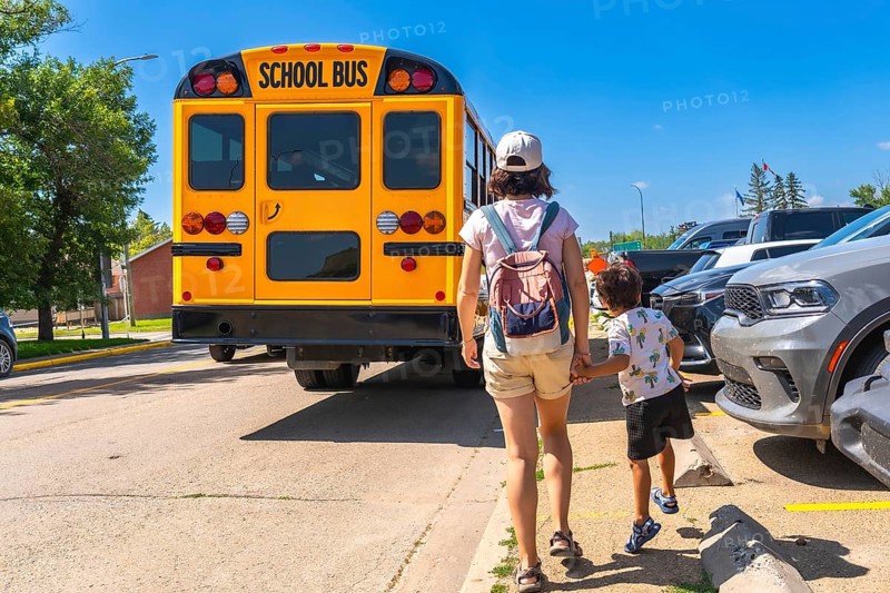 Mother walking her son to the school bus on the first day back to school, holding hands and heading towards the yellow bus parked near the school