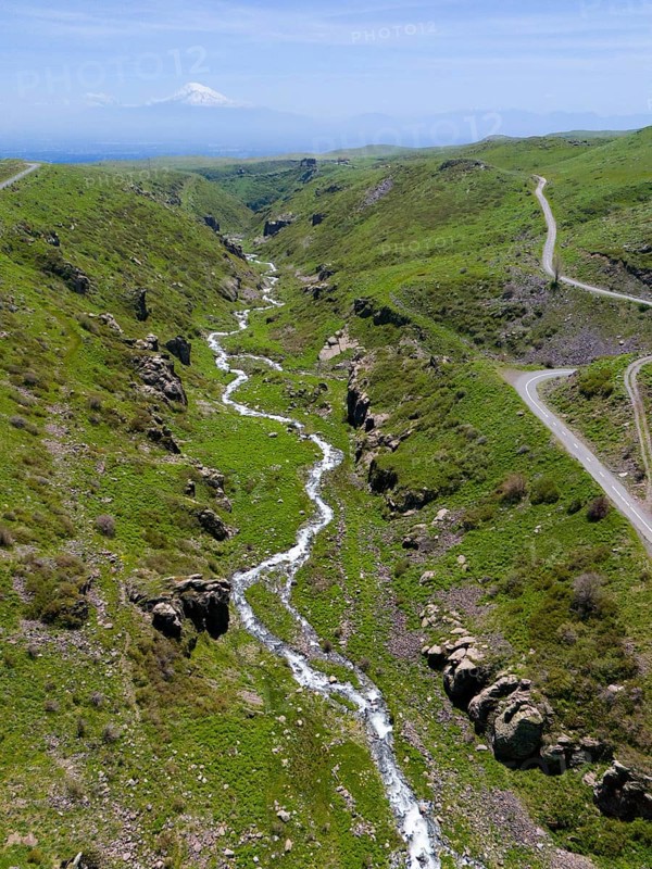 A panoramic view shows a river flowing through a green, hilly landscape, aerial view, river and gorge Arkashian, in the background Mount Ararat in Turkey, Aragazotn Province, Armenia
