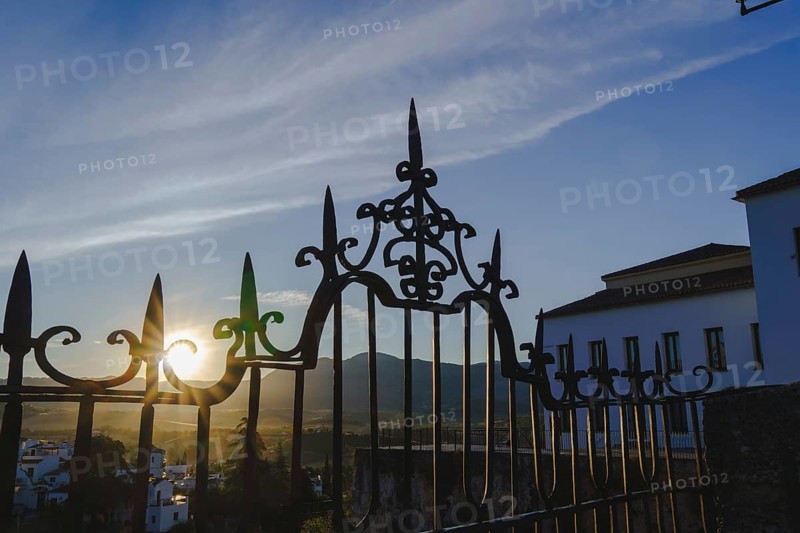 Sun shining through the ornate wrought iron gate at sunrise in ronda, spain, with mountains and white houses in the background in ronda, malaga, spain