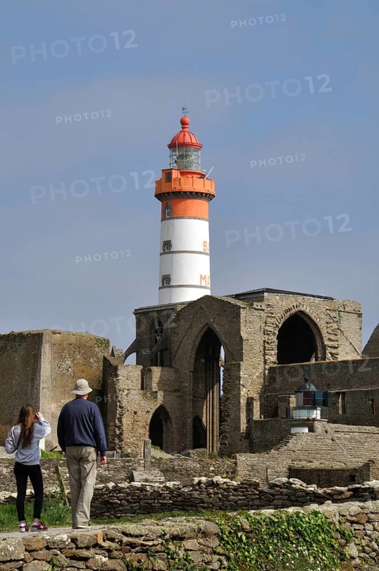 Pointe St Mathieu and Abbey Lighthouse, A historical lighthouse stands amidst ancient stone ruins under a clear blue sky, with people walking nearby in Brittany, France