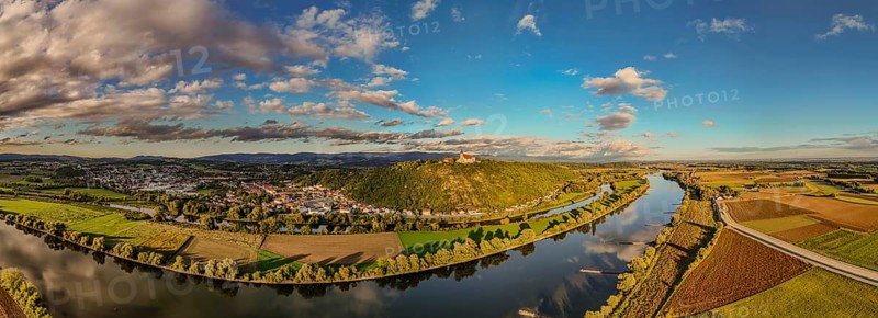 A picturesque landscape shows the Danube meandering through the green surroundings. Hills and clouds form an impressive backdrop at sunset. The pilgrimage church of the Assumption of the Virgin Mary on Bogenberg can be seen in the background. Bogen, Lower Bavaria