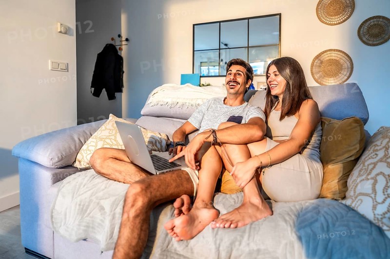 Young couple laughing while watching a streaming movie on their laptop, sitting comfortably on the sofa in their modern apartment, enjoying a relaxing evening together
