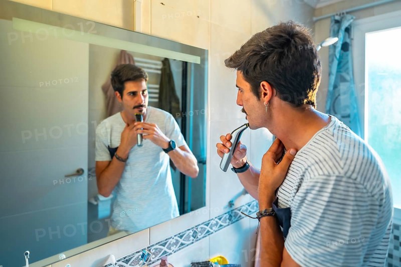 Caucasian man trimming his mustache with an electric razor while looking in the bathroom mirror during his morning routine