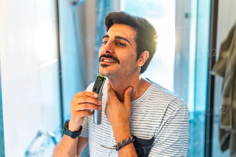 Man trimming his beard using an electric razor, taking care of his appearance in the bathroom mirror