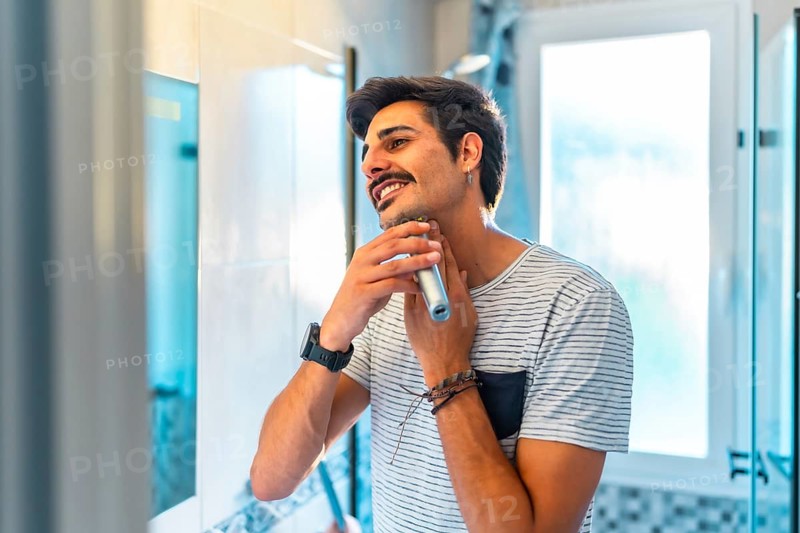 Happy young man trimming his beard in the bathroom mirror using an electric razor, enjoying his morning routine