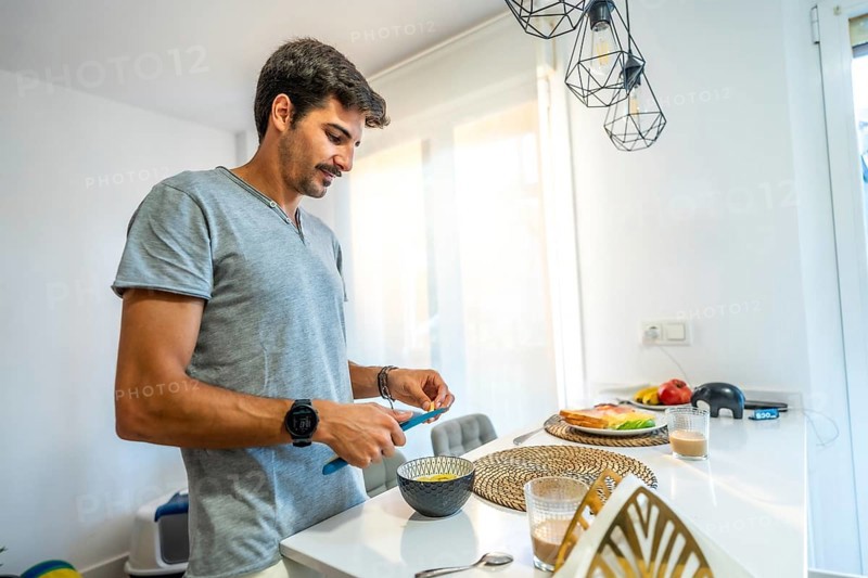 Young man spreading butter on fresh bread while enjoying breakfast in a bright, modern kitchen filled with natural light and comfort