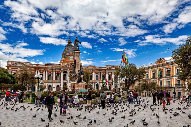 Main square full of people and pigeons in front of a large building with a statue under a blue sky, The presidential palace of La Paz in Bolivia
