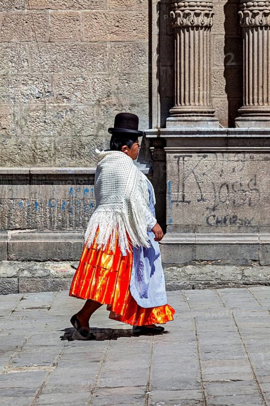 Woman in traditional dress with orange skirt walking along a historical wall, Traditionally dressed woman in the streets of La Paz Bolivia