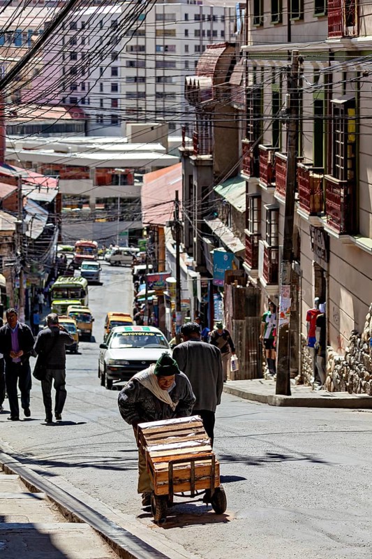 A man pushes a cart on a busy city street. Buildings and cars can be seen in the background, A man pushes a handcart through the streets of La Paz in Bolivia