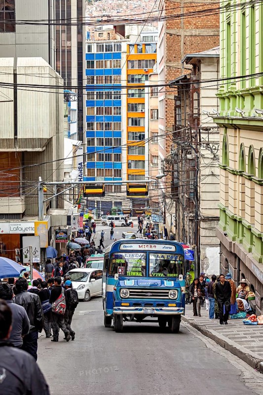 Busy city street with a blue bus and many people during the day in the middle of tall buildings, A colourful bus drives through the streets of La Paz in Bolivia