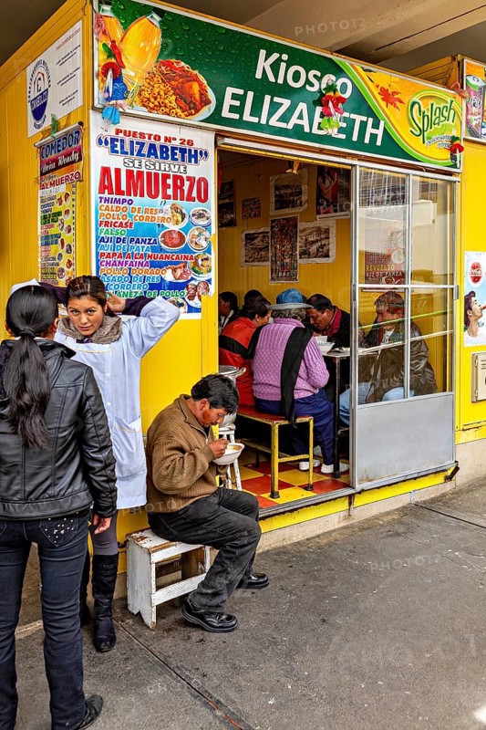 Small food stall with yellow walls, people sitting and eating, menu posters on the wall, Small shops and restaurants in the market halls of La Paz in Bolivia