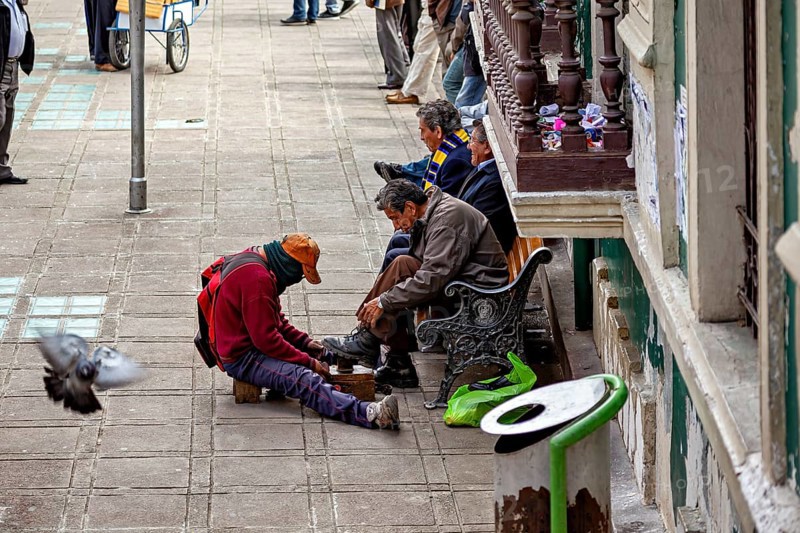 A shoeshine boy shines shoes on a street while passers-by and a pigeon are visible, shoeshine boy in the streets of La Paz in Bolivia