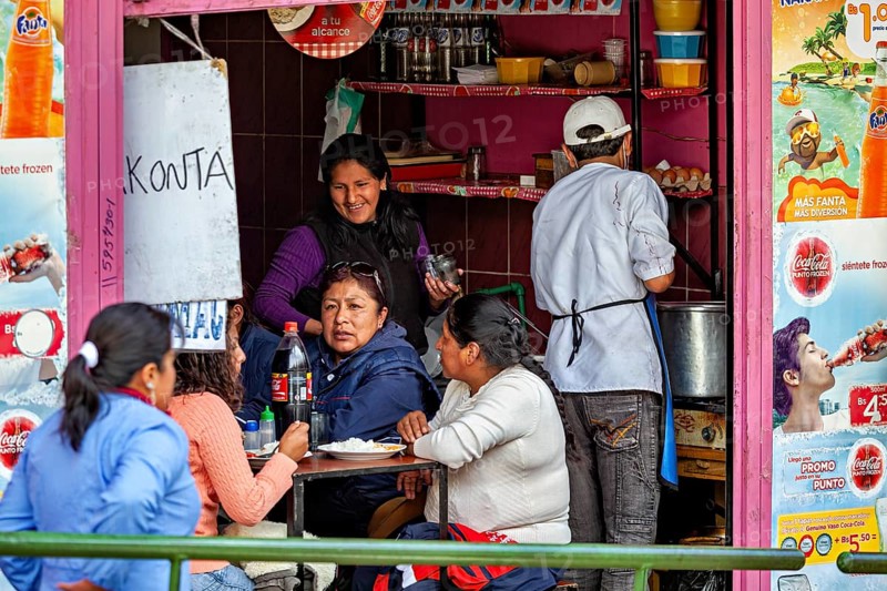 People enjoy their meals in the middle of a lively street snack bar, surrounded by colourful posters, small shops and restaurants in the market halls of La Paz in Bolivia