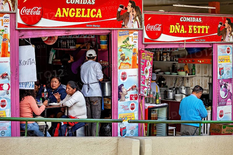A busy street snack bar with people sitting at tables and eating, surrounded by colourful advertising signs, Small shops and restaurants in the market halls of La Paz in Bolivia