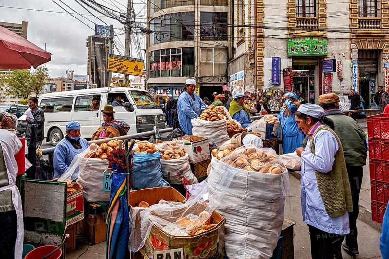 A bustling city market with bread stalls and vendors in a busy street between tall buildings, woman selling in the streets of La Paz, Bolivia