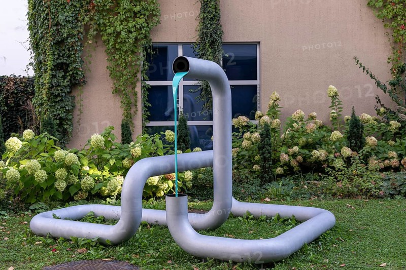 Metal sculpture in the garden with flowing coloured water and plants in the background, city centre Linz Upper Austria Austria