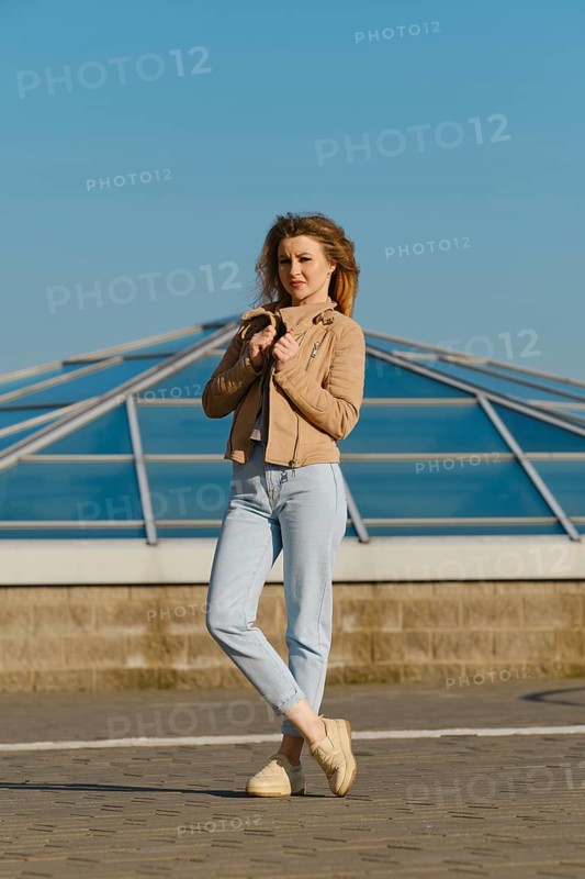 A young woman stands outdoors, wearing a light jacket and jeans. She holds her jacket gently while posing against a backdrop of a sleek glass structure under a clear blue sky