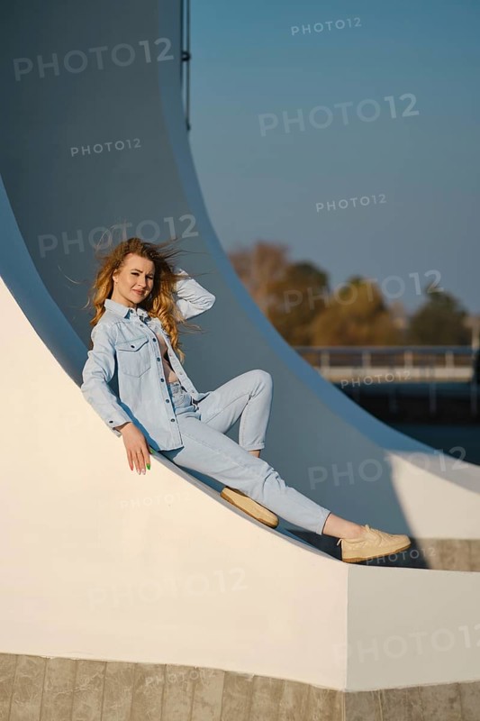 A woman with long hair is sitting comfortably on a curved architectural element. She wears a light denim outfit and smiles as sunlight highlights her pose