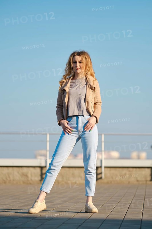 A woman stands on a rooftop during a bright sunny day. She is wearing a light fleece jacket and jeans, with her hair styled in loose waves. The clear blue sky provides a vibrant backdrop