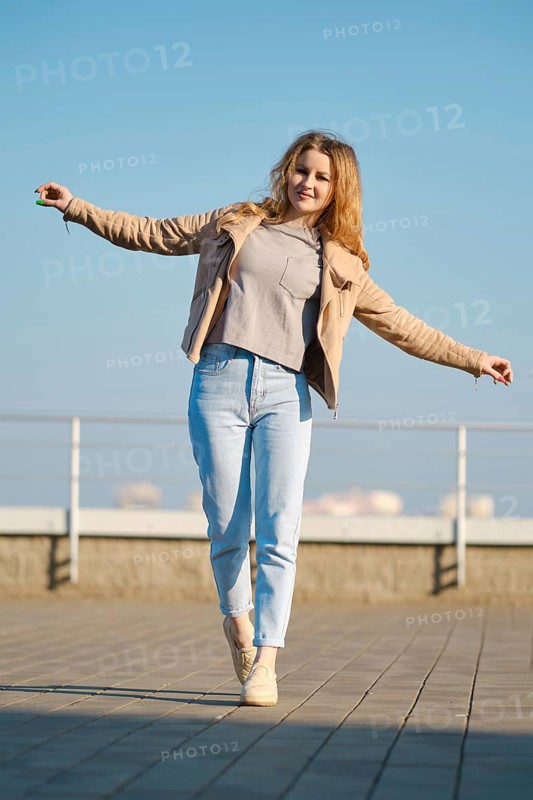 A young woman stands on one foot with arms outstretched, smiling under a clear blue sky. She wears a light fleece jacket and jeans, enjoying a sunny day at a city location with a railing in view