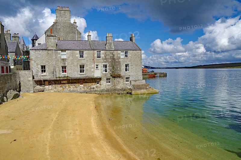 Old stone cottage on a quiet beach with clear water and partly cloudy skies, Lerwick, Shetland Islands, Scotland, United Kingdom