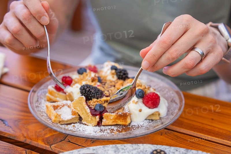 Hands holding spoons over waffles with cream and berries on a table