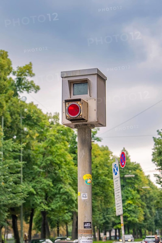 Traffic surveillance camera on a pole, surrounded by trees and road signs under a cloudy sky, Munich, Germany