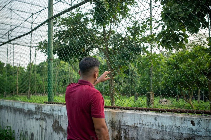 A man stands by a chain-link fence surrounded by lush greenery, appearing contemplative-Dhaka, Bangladesh