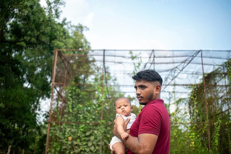 A man holds a baby outdoors by a metal structure with greenery in a relaxed setting-Dhaka, Bangladesh