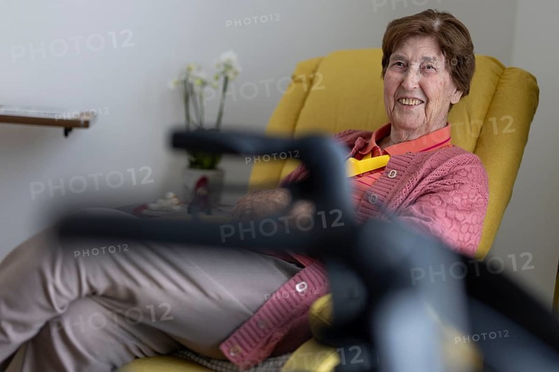 90+ year old senior citizen sitting laughing in her room in a retirement home behind a walker in a yellow armchair, Cologne, North Rhine-Westphalia, Germany