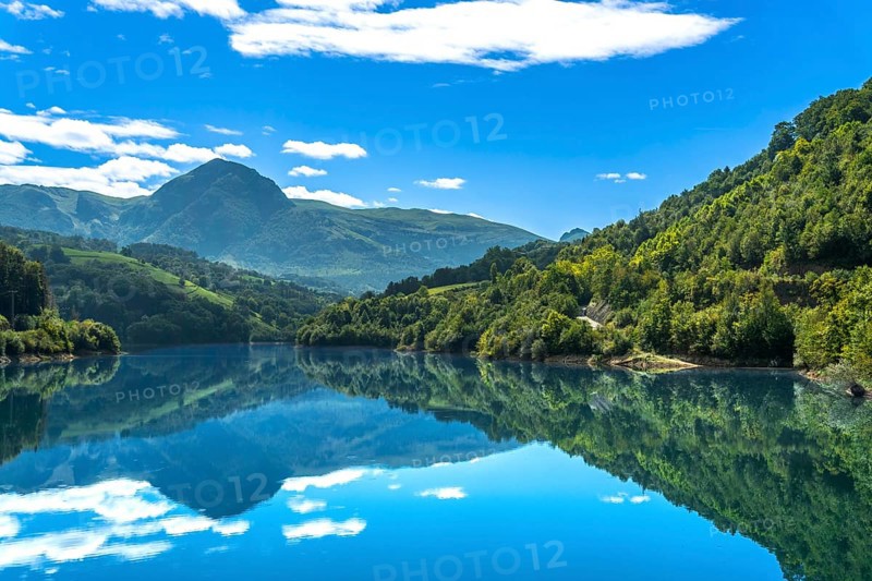The serene ibiur reservoir in tolosa, gipuzkoa, reflects the surrounding mountains and blue sky with white clouds on a sunny day, creating a tranquil scene in the heart of the basque country