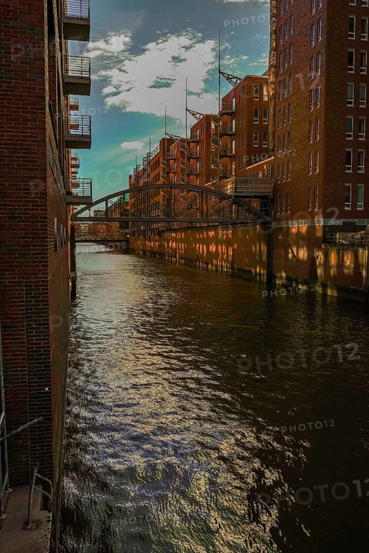 Sunlight is reflected in the water of the canal between tall brick buildings. View from the Wilhelminenbrücke over the Kehrwiederfleet towards the Speicherstadt, 20457 Hamburg, Germany