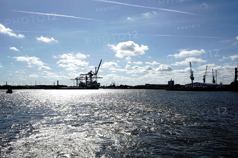 An industrial harbour with cranes and reflecting sunlight on the water under a blue sky. The view towards the Hansa harbour, seen from the Norderelbe in 20457 Hamburg