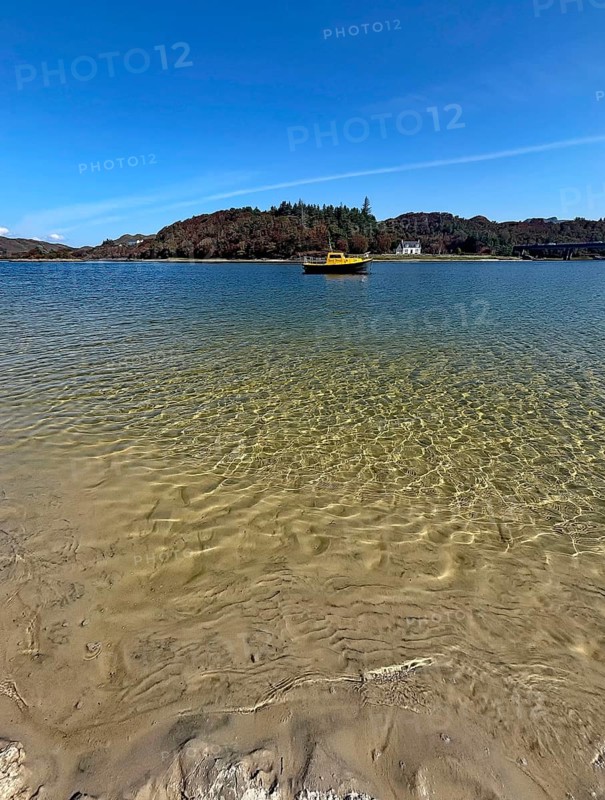 Clear water on a beach with a view of a yellow boat and wooded island in the background, Silver Sands of Morar, Morar, film set, Scotland, United Kingdom
